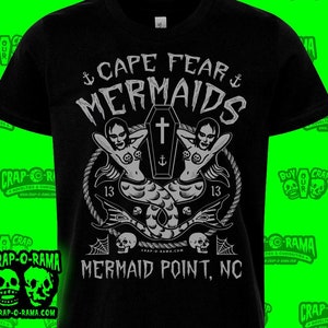 May include: Black t-shirt featuring a white graphic design of two mermaids flanking a coffin with a cross on top. The text "Cape Fear Mermaids" is above the image and "Mermaid Point, NC" is below the image.
