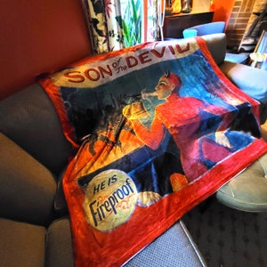 May include: Red and blue throw blanket with a vintage illustration of a devil figure with horns and a red suit. The text "Son of the Devil" and "He is Fireproof" are printed on the blanket.