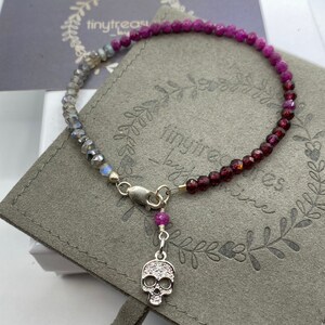 May include: A gradient gemstone bracelet with a silver skull charm. The bracelet features a mix of light blue and purple gemstones. The skull charm is adorned with clear crystals.