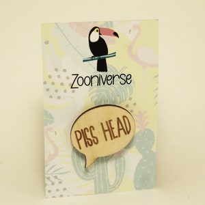 May include: A wooden speech bubble pin with the text "PISS HEAD" in brown lettering. The pin is on a card with a toucan and flamingo design and the text "ZooUniverse".