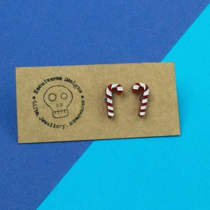 Puede incluir: Un par de pendientes de bastón de caramelo rojo y blanco en una tarjeta marrón con un logotipo de calavera que dice "Zooniverse Designs, Gifts, Jewellery, Homewares".
