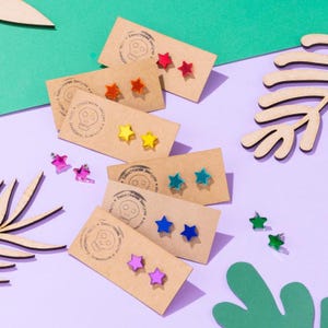 May include: A selection of star-shaped stud earrings in various colours, including red, orange, yellow, green, blue, and purple. The earrings are displayed on small, brown cards with a skull logo and text.