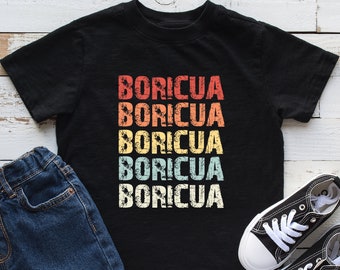 May include: Black t-shirt with the word "BORICUA" in a retro font, stacked vertically in red, orange, yellow, teal, and white. The shirt is paired with blue denim shorts and black and white sneakers.