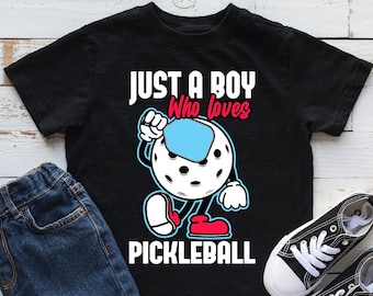 May include: Black t-shirt featuring a cartoon illustration of someone playing pickleball. The text "Just a boy who loves pickleball" is printed on the shirt.