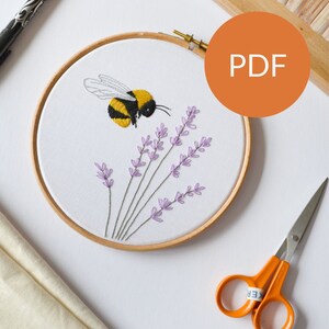 May include: Embroidery hoop with a detailed bee and lavender design. The bee is yellow and black, with lavender sprigs below. The hoop is made of wood, and the background is white. The image also includes scissors and a pen.