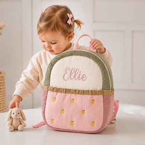 Puede incluir: Una mochila acolchada con una sección superior de color crema con el nombre Ellie bordado en letra rosa, un borde verde salvia y una sección inferior de color rosa empolvado decorada con un patrón de pequeñas frutas cítricas amarillas.
