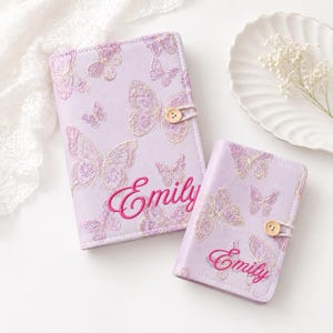 May include: Two light purple notebooks featuring embroidered butterfly patterns in white and pink thread. Each notebook has a wooden button closure and the name Emily embroidered in bright pink cursive script on the front cover.