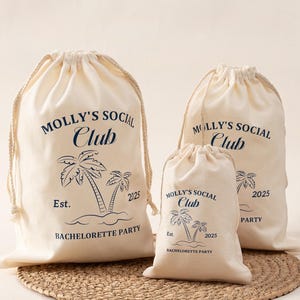 May include: Three beige drawstring bags with the text "MOLLY'S SOCIAL CLUB" and a palm tree design. A bottle of red wine and a champagne flute are next to the bags. The bags are on a round woven mat.