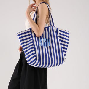 Puede incluir: Un bolso tote grande con un patrón de rayas verticales azules y blancas. El bolso tiene dos correas para el hombro y está bordado con las letras SGH en una fuente azul claro en el centro frontal. La tela presenta una textura tejida visible.