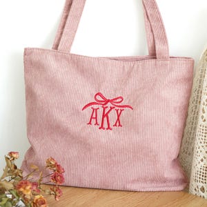 May include: A pink corduroy tote bag with long handles. The bag features an embroidered red monogram of the letters AKX. The bag is sitting on a wooden surface with dried flowers in the foreground.