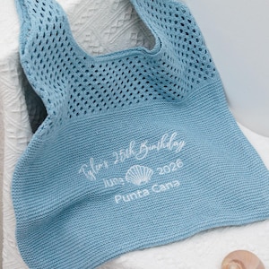 Puede incluir: Un bolso tote tejido de color azul claro con un asa grande. El bolso tiene un diseño similar a una malla en la mitad superior y tejido liso en la parte inferior. El texto en el bolso dice "Tyler's 25th Birthday June 2026 Punta Cana". Una concha marina descansa sobre la superficie blanca.