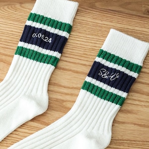 Può includere: Due paia di calzini bianchi con strisce verdi e blu scuro. Un paio ha il testo "0.06.24" e l'altro paio ha il testo "S&U".