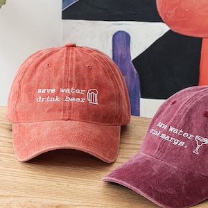 Può includere: Due cappellini da baseball rossi e bordeaux con testo ricamato. Il cappello rosso dice "save water drink beer" con un'icona di boccale di birra. Il cappello bordeaux dice "save water drink margs." con un'icona di bicchiere di margarita.