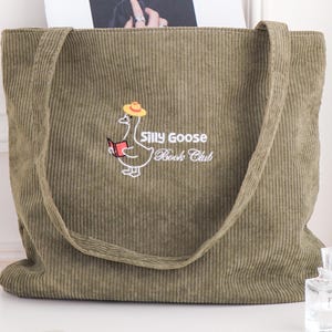 May include: Olive green corduroy tote bag with long straps. The bag features an embroidered cartoon goose wearing a hat and holding a book, with the text "Silly Goose Book Club". A perfume bottle and a phone are on the table.