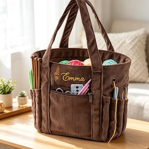 Peut inclure: Un sac fourre-tout de loisirs créatifs en velours côtelé marron avec le prénom Emma brodé et un petit motif floral. Le sac possède plusieurs poches extérieures contenant des aiguilles à tricoter et des crochets, avec des pelotes de laine colorées.