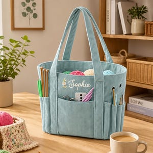 May include: A light blue corduroy craft tote bag featuring the embroidered name Sophia and a small floral accent. The bag has multiple exterior pockets filled with various knitting needles and crochet hooks, with a structured open-top design.