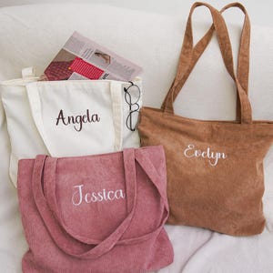 Puede incluir: Tres bolsos tote de pana en crema, rosa empolvado y marrón. Cada bolso tiene un nombre bordado en blanco: "Angela", "Jessica" y "Evelyn". El bolso crema contiene una revista y gafas. Los bolsos tienen asas largas.