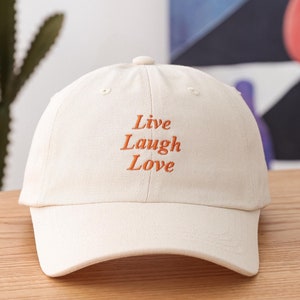 Peut inclure: Une casquette de baseball blanche avec un message brodé orange qui dit "Live Laugh Love".