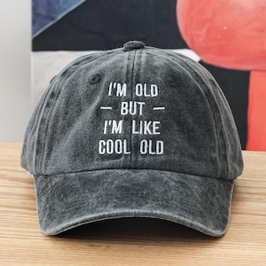 K&ouml;nnte beinhalten: Eine schwarze Baseballkappe mit wei&szlig; gesticktem Text, der "I'm old - but - I'm like cool old" lautet.