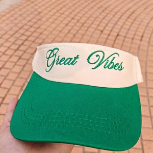 May include: A green and cream visor with the embroidered text "Great Vibes" in green script. The visor features a curved brim, designed to shield the eyes from the sun. The visor is made of a fabric material.