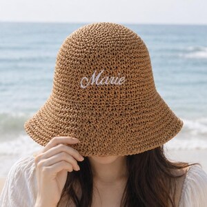 May include: A woven straw sun hat with a wide brim, embroidered with the name "Marie" in white script. The hat is a light brown color and is being held up by a person on a beach with the ocean in the background.