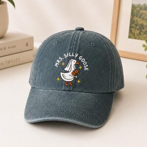 May include: A dark gray denim baseball cap featuring a white graphic of a goose wearing a veil and holding orange flowers. The text "MRS. SILLY GOOSE" is printed in a white arc above the goose, accented by small yellow four-pointed star icons.