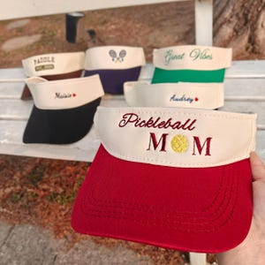 Può includere: Una collezione di visiere con vari disegni e testo. Una visiera è rossa e bianca con "Pickleball MOM" ricamato sul davanti. Altre visiere includono "Paddle", "Maisie", "Audrey" e "Great Vibes".