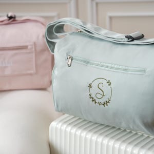 May include: A light blue duffel bag with a zippered pocket and an embroidered green wreath with the letter "S" in the center. The bag is sitting on a white surface.