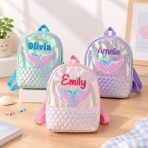 Puede incluir: Tres mini mochilas iridiscentes con un gráfico de cola de sirena y nombres bordados. Las mochilas son de color azul claro, morado claro y rosa claro, cada una con un patrón de escamas texturizado en el bolsillo inferior y correas a juego.