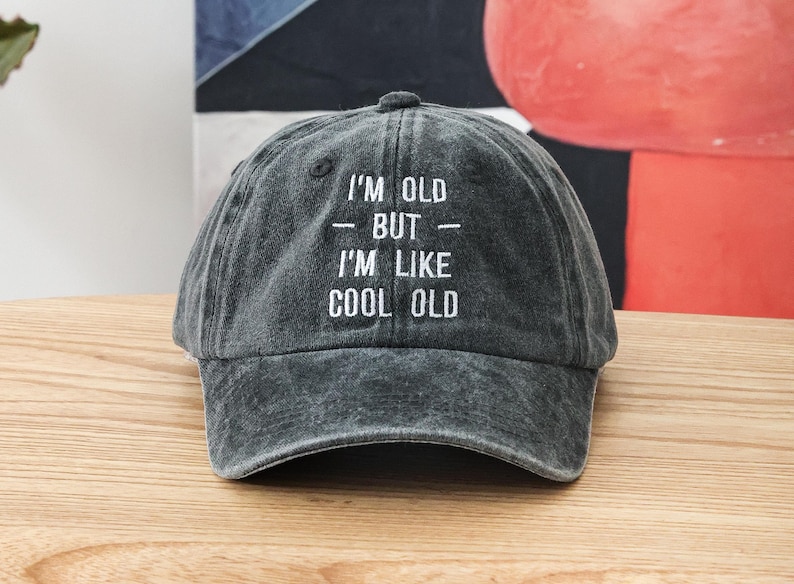 K&ouml;nnte beinhalten: Eine schwarze Baseballkappe mit wei&szlig; gesticktem Text, der "I'm old - but - I'm like cool old" lautet.