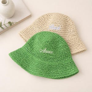 May include: Two woven bucket hats. One is green with the name "Anna" embroidered in white. The other is beige with the name "Bria" embroidered in blue. Both hats have a wide brim and a textured appearance.