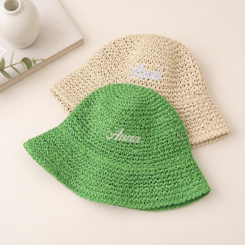 May include: Two woven bucket hats. One is green with the name "Anna" embroidered in white. The other is beige with the name "Bria" embroidered in blue. Both hats have a wide brim and a textured appearance.