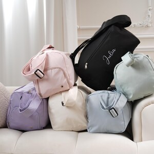May include: Five different colored duffel bags, including a black bag with the name "Julia" embroidered on it. The bags are made of a soft, canvas-like material and have a zipper closure.