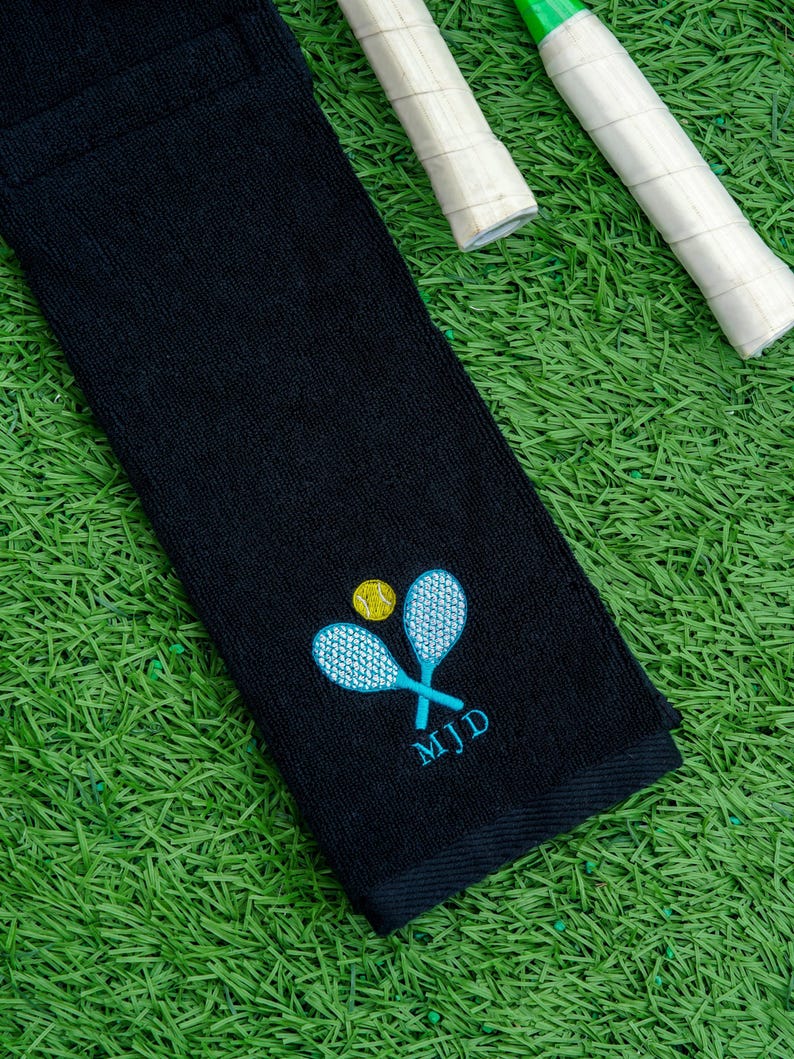 May include: Black tennis towel featuring embroidered tennis rackets, a yellow tennis ball, and the initials MJD in teal. Two tennis racket handles are visible, set against green artificial turf.