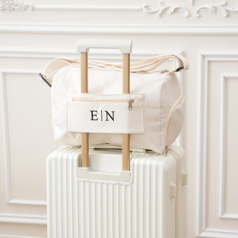 May include: A white canvas duffel bag with a zippered pocket on the front. The pocket has the letters "E|N" embroidered in black. The bag is sitting on top of a white suitcase with a handle.