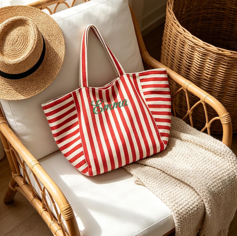 May include: A canvas tote bag featuring vertical red and white stripes with the name Emma embroidered in green cursive script on the front. The bag has two matching striped shoulder straps and a spacious open top design for carrying items.