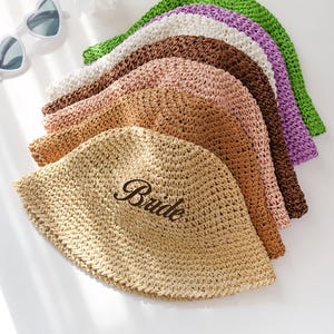 May include: A collection of woven straw hats in various colors, including beige, brown, pink, white, purple, and green. One hat is embroidered with the word "Bride". Heart-shaped sunglasses and a flower are in the background.