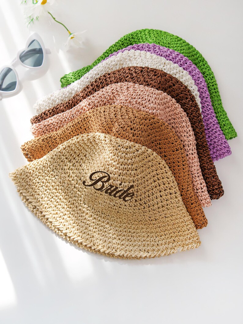 May include: A collection of woven straw hats in various colors, including beige, brown, pink, white, purple, and green. One hat is embroidered with the word "Bride". Heart-shaped sunglasses and a flower are in the background.