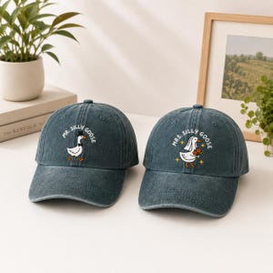 Puede incluir: Dos gorras de béisbol de mezclilla azul oscuro con gráficos de gansos bordados. La gorra izquierda muestra un ganso con sombrero de copa y pajarita con el texto MR. SILLY GOOSE. La derecha muestra un ganso con velo y flores con MRS. SILLY GOOSE.