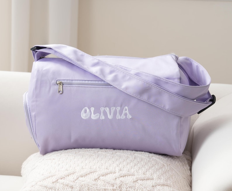 May include: A lavender-colored duffel bag with a zippered pocket and a white embroidered name "Olivia".