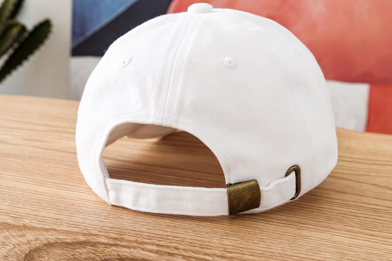 K&ouml;nnte beinhalten: Eine wei&szlig;e Baseballkappe mit einem braunen verstellbaren Riemen. Die Kappe ist auf links gedreht und zeigt die R&uuml;ckseite der Kappe.