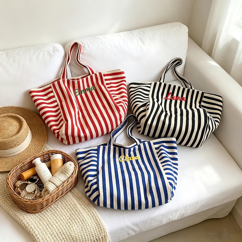 May include: Three canvas tote bags with vertical stripes in red, black, and blue. Each bag features two handles and an embroidered name on the front: Emma in green, Luna in red, and Clara in yellow. A straw hat and a wicker basket with items sit nearby.
