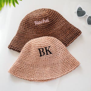 May include: Two woven straw bucket hats. One is brown with "Rachel" embroidered in pink. The other is tan with "BK" embroidered in black. White heart-shaped sunglasses are in the background.