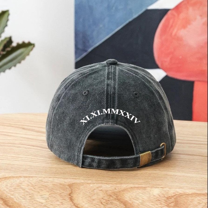 K&ouml;nnte beinhalten: Eine schwarze Baseballkappe aus Denim mit verstellbarem Riemen. Die R&uuml;ckseite der Kappe ist mit wei&szlig;er Stickerei versehen, die "XI.XI.MMXXIV" lautet.