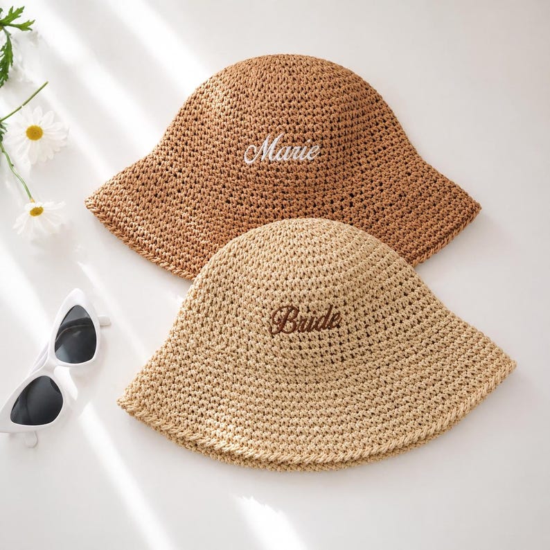 May include: Two straw sun hats. One is light brown with the name "Marie" embroidered in white. The other is beige with the word "Bride" embroidered in brown. Both hats have a wide brim and are made of woven straw.