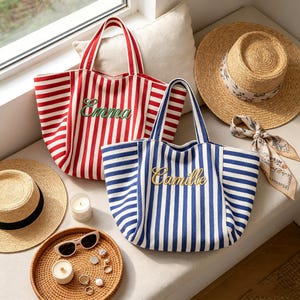 May include: Two canvas tote bags with vertical stripes, one in red and white with the name Emma embroidered in green, and one in blue and white with the name Camille embroidered in gold. Two straw hats, sunglasses, and small candles are nearby.