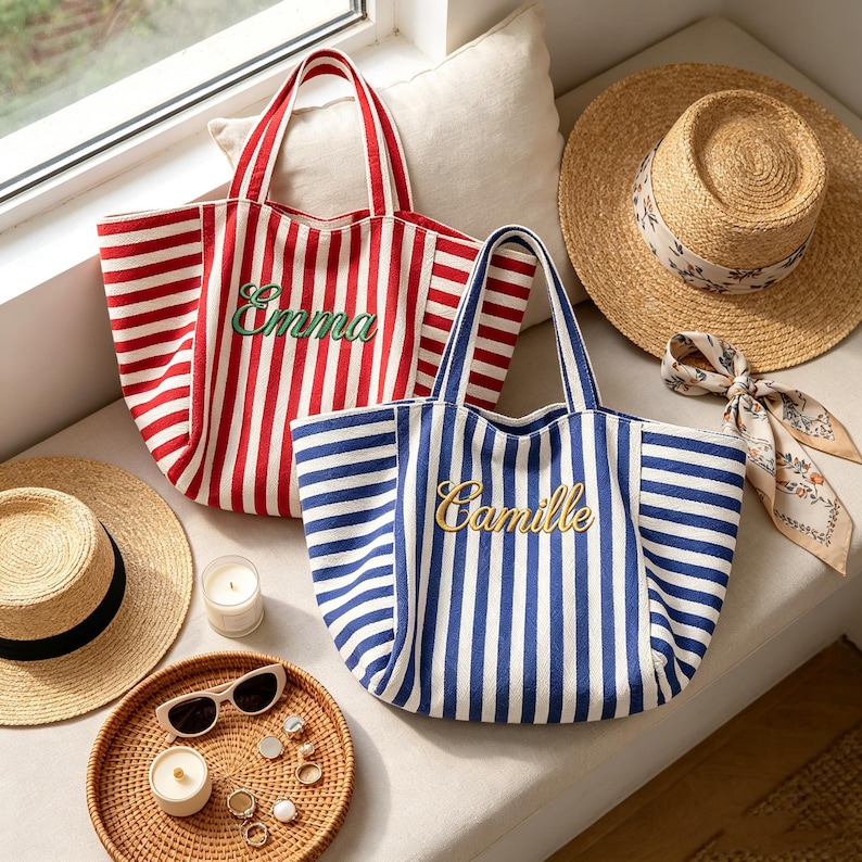 May include: Two canvas tote bags with vertical stripes, one in red and white with the name Emma embroidered in green, and one in blue and white with the name Camille embroidered in gold. Two straw hats, sunglasses, and small candles are nearby.