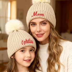 May include: Two beige knit beanies with cream-colored faux fur pom-poms. One beanie has the word "Mom" embroidered in red, the other says "Ophelia". The hats are worn by a mother and daughter.