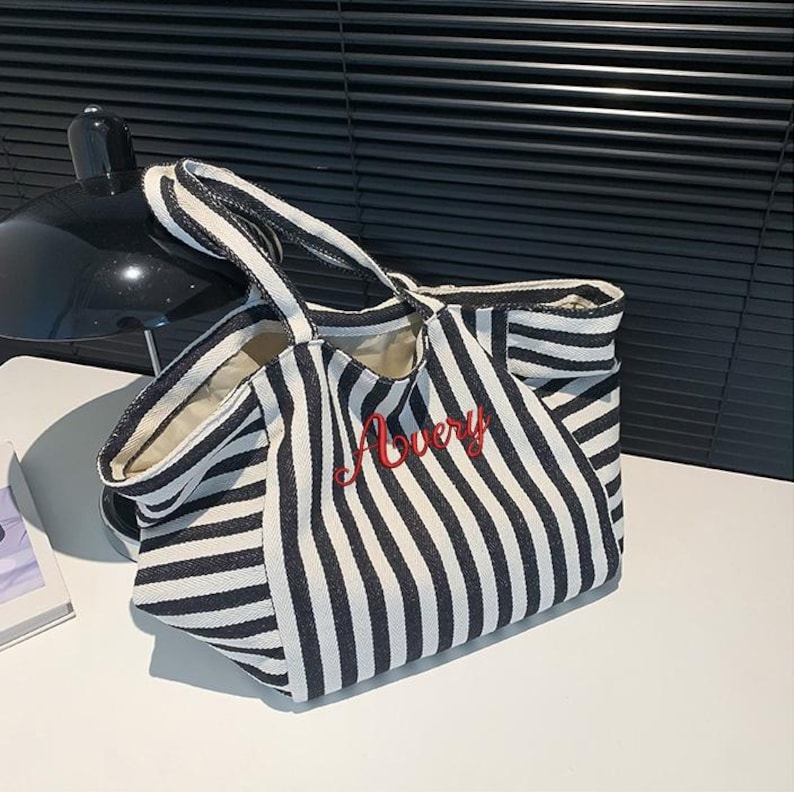 May include: A black and white striped canvas tote bag featuring two top handles and the name Avery embroidered in red cursive script on the front. The bag has a structured base and an open top design with a light-colored interior lining.