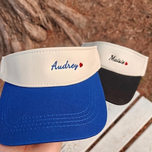 Può includere: Due visiere, una blu e una nera, con fasce color crema. La visiera blu ha "Audrey" ricamato in blu con un cuore rosso, e la visiera nera ha "Maisie" ricamato in nero con un cuore rosso. Entrambe le visiere sono per la protezione solare.